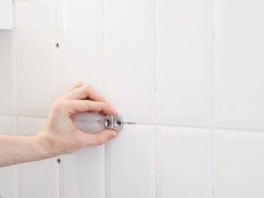 Genç bir adam, tanınmaz halde, eliyle banyodaki beyaz fayanslı duvardaki havlunun metal bir parçasını çıkarıyor, yakın çekimde..