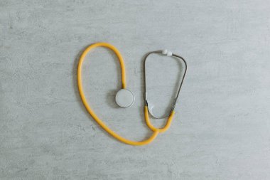 Bir sarı steteskop, merkezde açık gri bir taş zemin üzerinde kalp şeklinde, düz yakın plan yatmakta. Sağlık konsepti, sağlık günü, tıbbi gereçler.