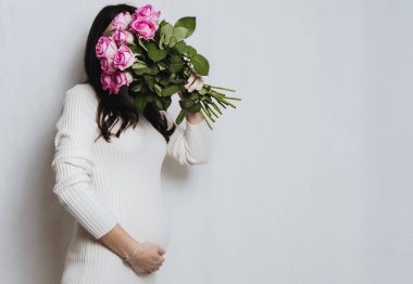 Beyaz örgü elbiseli genç esmer bir kadın beyaz bir duvarın sol tarafında duruyor, bir eliyle yuvarlak göbeğini tutuyor ve diğer eliyle yüzünü pembe güllerle kaplıyor. Sağ tarafta fotokopi alanı olan yakın çekim kenarı. Kavram