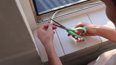 Beyaz tenli genç, kendine yapışkan beyaz kauçuk profili yeşil makasla kesiyor ve pencere çerçevelerine yapıştırıyor. Yan tarafından yavaş çekimle yakın çekim. Pencere çerçevelerinin onarımı ve yalıtımı kavramı, bahar çalışması.