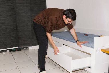 Siyah saçlı beyaz bir adamın portresi. Beyaz bir yatağın üzerine eğilip çekmeceyi çekiyor. Akşamları salonda toplandıktan sonra kontrol ediyor. Yatak kurulumu kavramını, adım adım talimatları.