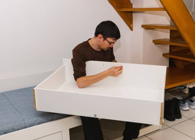 Genç bir Kafkas esmer adam ağzında bir vida tutuyor ve geceleyin merdivenlerin altında oturma odasında otururken bir yatak çerçevesini monte ederken onu parmaklarıyla elle sıkıyor. Orta ölçekli yan görünüm.