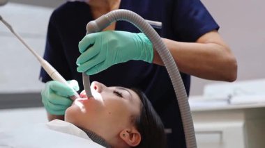 Mavi üniformalı genç bir beyaz kadın dişçi ve tartar ve plaketten dişleri temizleyen yeşil eldivenler. Hasta hasta için özel aletler var. Kapalı, yakın plan, alt taraf manzaralı. Sakız ve diş tedavisi kavramı, dişçi günü.
