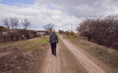 Bulutlu bir sonbahar gününde kırsalda toprak bir yolda yürüyen, kıdemli, sarı saçlı bir kadının yakın plan görüntüsü. Kavram: Emeklilik yaşam tarzı.
