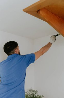Beyaz tenli genç bir esmer adam mavi bir tişörtle arkada duruyor ve duvarların köşelerini beyaz boyayla boyuyor. Tahta bir merdivenin altında küçük bir fırça, yakın plan yan görüş..