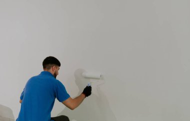 Mavi tişörtlü, koyu renk sakallı, genç bir beyaz adam arkadan yarım yamalak oturuyor ve beyaz boyalı bir duvar boyuyor, yakın çekimde seçici odak noktası ve sağ tarafta kopyalama alanı var..