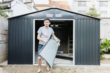 Bir yaz günü arka bahçeye yerleştirmeden önce metal bir barakadan çelik kapı panelini çıkaran beyaz bir işçinin portresi. Profil dökümü oluşturma kavramı.