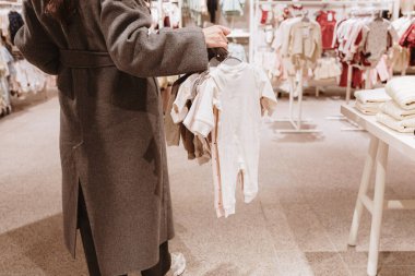 Gri paltolu genç, tanınmayan beyaz bir kadın alışveriş merkezinden alışveriş yapmak için kasaya doğru yürürken askılarda çeşitli yeni doğan bebek kıyafetleri taşıyor. Orta boy, yan görüş. Konsept: Çevrimdışı alışveriş zamanı.