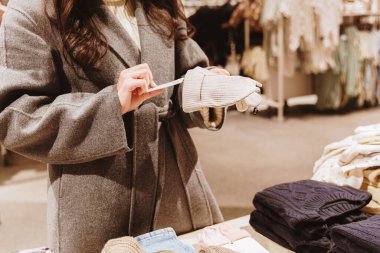 Gri paltolu, genç, tanınmayan beyaz bir kadın, kulaklı, kurdeleli bir bebek şapkası tutuyor ve alışveriş merkezinde alışveriş yaparken kağıt fiyat etiketini inceliyor. Yakın çekim, yukarıdan aşağıya manzara. Konsept: Çevrimdışı alışveriş zamanı, kıyafet seçme.