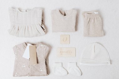 Güzel, yumuşak örülmüş ve yeni doğmuş pamuklu bebek kıyafetleri krem rengi ve bej renginde, üzerinde 0 ay yazılı tahta tabelalar hafif taş zemin, düz zemin ve yakın çekim şeklinde katlanır. Çocuk kıyafetleri, çocuk büyümesi, hediye setleri.