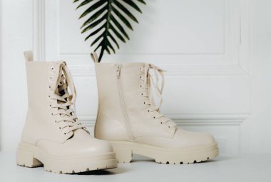 Bir çift şık krem rengi kadın çizmesi, kalın tabanlı, bağcıklı ve yan kapaklı, palmiye dallı bir kapının yanında duruyor. Yan görüntüyü yakından çek. Kadın ayakkabıları konsepti, moda koleksiyonu.