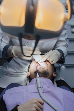 Bir dişçi klinikte sakallı bir genç için diş içi tarayıcı kullanarak sol azı dişlerinin dijital üç boyutlu modelini yapar. Yukarıdan ve yandan dikey olarak yaklaş. Konsept: Diş sağlığı, teknoloji kullanımı, dişçiler günü.