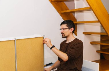 Gözlük takan genç esmer bir adam, bir yatak monte ederken çekmecenin kenarına metal bir örtü takar, akşam vakti bir odanın altında, orta ölçekli, kenar manzaralı bir odada oturur. Kavramsal yatak montajı.