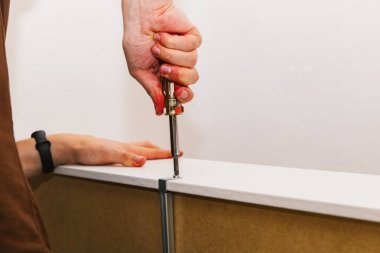 Genç bir zanaatkar, akşam bir odada bir yatağın montajı sırasında çekmeceye metal bir raptiye bağlayan yuvarlak bir minifiks takmak için tornavida kullanır. Kavramsal yatak montajı.