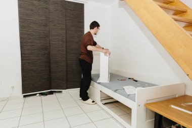 Gündelik giysiler içinde genç bir işçi, akşamları bir odaya yatak yerleştirirken manüel olarak bir kutu monte eder. Kavramsal yatak montajı.