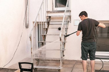 Genç beyaz adamın portresi kirli metal merdiveni yıkamak uzun yüksek basınçlı su jeti ile arka bahçede duran basınçlı yıkama makinesini kullanmak bahar günü, yan görüş. Bahar zamanı konsepti teknolojiyle çalışır..
