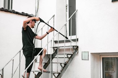 Beyaz tenli bir adamın portresi, koyu renk giysiler içinde çıplak ayakla, metal merdivenlerin alt kısmını yıkarken, basınç altında uçan bir su püskürtüsüyle evinin arka bahçesindeki kercher 'ın yardımıyla,