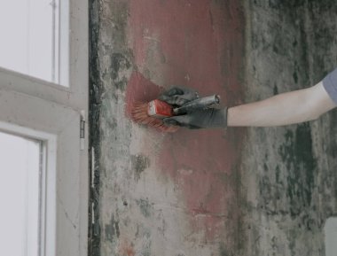 Eldivenli tanınmayan genç bir adam kirli bir duvara fırçayla kırmızı çimento sürüyor. Küflü lekeler, yakın çekim. Ev tadilatı ve inşaat işi kavramı.