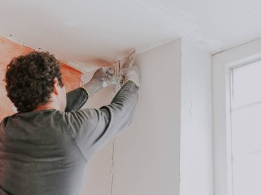 Üniformalı, kıvırcık saçlı, genç bir beyaz adam sıva ve inşaat eldivenleri, pencerenin yakınındaki duvardan bir inşaat feneri çekmek için bir spatula kullanıyor. Daire yenileme konsepti.