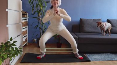 Noel Baba şapkası ve Noel çorapları giyen genç beyaz bir kadın, paspasın üzerinde elastik bir bant ve kanepede bir köpekle çömelme egzersizi yapıyor. Ortalama çekim, gerçek hareketle yan görüş. Ev fitness konsepti.