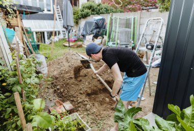 Siyah tişörtlü ve mavi şortlu genç bir beyaz işçi, bir yaz günü arka bahçedeki yığının üzerine bulanık bir şekilde düşen kürekle toprağı hareket ettiriyor. Yan görüş, orta çekim, alan derinliği. Kavram: kazı çalışması zamanı.