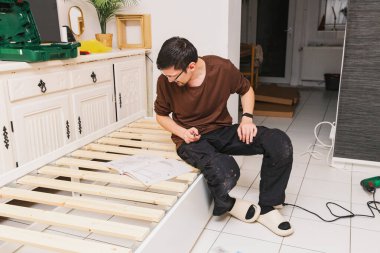 Yakışıklı, genç, beyaz bir işçi, geceleri odasında otururken bir yatak kurma talimatlarını dikkatle inceler. Orta boy, yan görüş. Kavramsal yatak montajı.