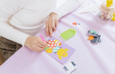 Tanımlanamayan beyaz bir kızın portresi, bir masada oturuyor ve el yapımı bir kart hazırlıyor bir bahar günü evde, mutfakta paskalya tavuğuna yapay elmas yapıştırarak, yan tarafından üst manzara, yakın çekim. Yaratıcılığı kendi yaratıcılığınla kavra.