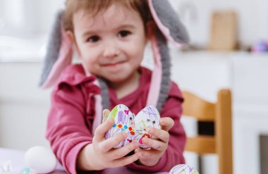 Gri tavşan bantlı, elinde keçeli kalemle süslenmiş iki DIY Paskalya yumurtası tutan beyaz saçlı güzel bir kız bir bahar günü masada oturuyor, tarlanın derinliği ile kenar manzaralı. Mutlu Paskalyalar, DIY