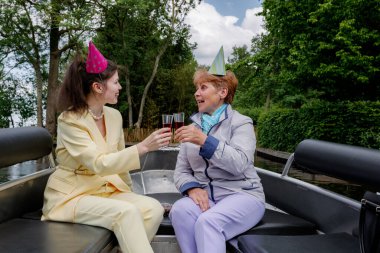 Tereyağından kıl çeker gibi mutlu genç bir kızın portresi ve kırmızı içkiyle bardakları tokuşturup doğum gününü kutlayan ve eğlenen olgun bir annenin portresi. Bahar günü tekne kanalında yelken açmak, yan görüş, orta çekim. Yat.