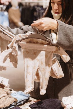 Gri ceketli, güzel, beyaz tenli bir kadın alışveriş merkezinde alışveriş yaparken askıda duran iki yeni doğmuş bebek kıyafetinden birini tutuyor ve inceliyor. Yakın plan, yan görüş. Konsept: Çevrimdışı alışveriş zamanı, kıyafet seçme.