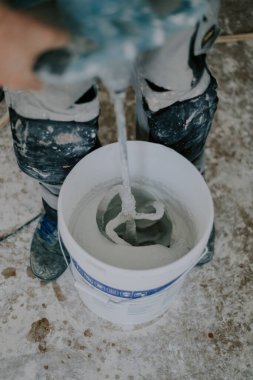 Beyaz tenli, tanınmaz halde beyaz macunu beyaz bir kovada, küçük bir inşaat mikseriyle kaplayan, kirli bir evin içinde duran, üst tarafı seçici bir odak ile yakın çekim yapan genç bir erkek. Ev yenileme konsepti, inşaat işi.