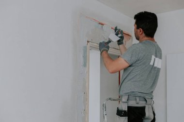 Gri üniformalı genç bir beyaz tenli esmer, eldivenlerindeki spatulayla bir inşaat alanını temizliyor, kapının köşesindeki merdivende yarı açık duruyor, aşağıdan seçici bir odak ile yakın çekim yapıyor. Şey...