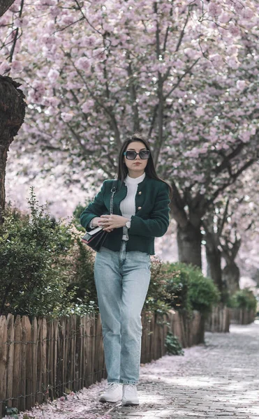 Beyaz saçlı, yeşil ceketli ve güneş gözlüklü güzel bir esmerin portresi bir bahar günü çiçek açan sakura ağaçlarıyla dolu bir şehir caddesinde dikiliyor..
