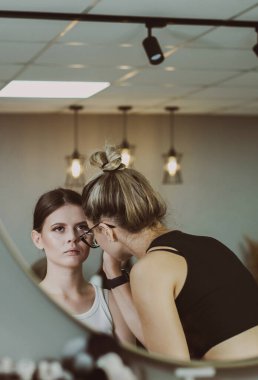 Genç ve güzel bir beyaz makyaj sanatçısı, bir sandalyede yan yana oturan bir kızın sağ göz kapağına göz farı sürüyor ve sabahın erken saatlerinde, bir güzellik salonunda, saha derinliği ile yakın bir yan görünüm yansıtıyor. Adım