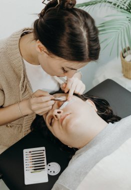 Beyaz tenli güzel bir kızın portresi, kozmetik bilimci yapay kirpikleri kadın bir müşterinin sol gözüne yapıştırıyor. Kozmetoloji masasında bir bahar günü, üst taraftaki yakın çekim manzarasında yatıyor. Kirpik.