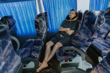 Gri takım elbiseli, boynunda yastıkla, çıplak ayaklarıyla koridora doğru uzanan, uzun bir yolculuktan yorgun, yorgun, mavi perdeli, yol boyunca ilerleyen bir otobüste oturan, beyaz saçlı, yakışıklı bir genç.