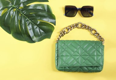 Büyük altın zincirli, güneş gözlüklü ve sarı arka planda sarı yapraklı, sağda küçük bir fotokopi alanı bulunan şık, yeşil örtülü çanta. Modaya uygun aksesuarlar, seyahat, pankart.
