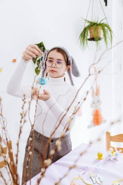 Beyaz tenli güzel bir kızın portresi masada duruyor ve söğüt dalına mavi kağıt asıyor, bahar günü mutfakta Paskalya buketi yapıyor, orta ölçekli. Mutlu çocukluk kavramı, mutlu paskalyalar, çocuk