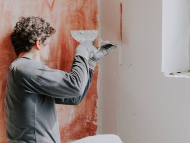 Üniformalı, kıvırcık saçlı, genç bir beyaz adam sıva ve inşaat eldivenleri, pencerenin yakınındaki duvardaki inşaat fenerini kapamak için spatula ve macun kullanıyor. Daire yenileme konsepti.