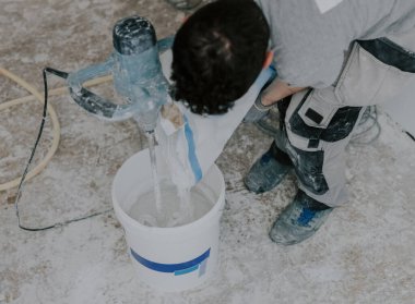 Genç bir beyaz adam, yenilemenin devam ettiği odada ayakta duruyor ve bir torbadan kuru macunu bir kovaya doldurup, bir inşaat mikseriyle karıştırıyor, seçici odaklı orta ölçekli bir yan görüş kullanıyor. Ev yenileme konsepti