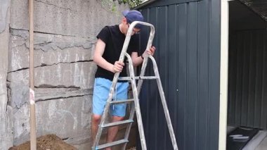 Genç bir beyaz zanaatkar, bir yaz günü arka bahçedeki metal bir barakanın çatısını kurmak için merdivene tırmanır. Yan görüş, orta çekim, ağır çekim. Kavram: Metal döşeme, çatı kurulumu.