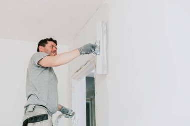 Genç, mutlu, beyaz bir adam merdivenin üzerinde duruyor ve kapının üstündeki duvarı bahar günü büyük bir spatulayla kaplıyor. Yan taraftan seçici bir odak ile yakın plan görüntüsü. Sıva ve inşaat işi kavramı.