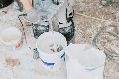 Çalışan üniformalı, beyaz ıslak macunu beyaz kovayla kırbaçlayan, beyaz teneke giyen, odada duran, üst taraftan seçici odaklı, tanınmayan beyaz bir genç. Ev yenileme konsepti, inşaat işi.