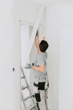Üniformalı, olgun, beyaz bir işçi merdivenin yanlamasına ayakta duruyor ve geniş bir malayla kapının köşesine duvar yapıştırıyor. Dikey yan görüş, yakın plan. Konsept: Ev yenileme, inşaat işi, kapı bitirme.