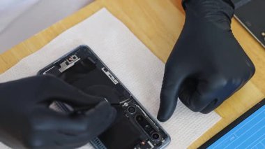 Tanımlanamayan siyah lateks eldivenli genç bir beyaz adam özel cımbız kullanarak akıllı bir telefona mini bir cıvata yerleştirir, tamir eder, bir yaz günü mutfakta masada oturur, üst tarafı yavaş çekimde çok yakından görür. Cep telefonu