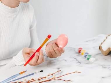 Beyaz balıkçı yaka kazaklı beyaz bir genç kızın elleri, paskalya bayramına hazırlanmak için mermer bir masada otururken elinde pastel pembe akrilik fırçayla resim yapıyor. El işi, dikiş işi kavramı.