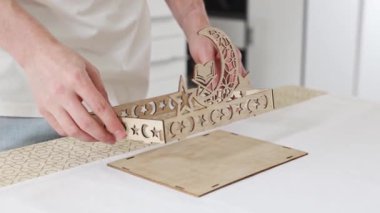 Masada duran genç beyaz bir adam dipteki deliklere dört adet yan parça yerleştiriyor, oyulmuş ahşap bir tepsi yapıyor. Yakın çekim, yavaş çekimde yan görüş. Konsept: tepsi montajı, Ramazan Kareem, Bayram Mübarek.