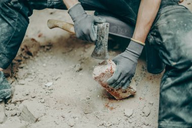 İş kıyafetleri içinde tanınmayan genç bir beyaz adam, balta kullanarak toprak ve eski çimentodan tuğlaları temizliyor, küçük bir sandalyeye oturuyor, bacakları açık bir şekilde onarımların devam ettiği bir odaya yayılıyor, yakından bakıyor. İnşaat işi kavramı.