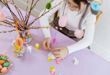 Portrait of one beautiful Caucasian girl in bunny headband preparing felt stickers to stick to pussy willow branch with hanging toys, creating Easter bouquet on spring day, top side view close-up selective focus. Concept of happy childhood, happy eas