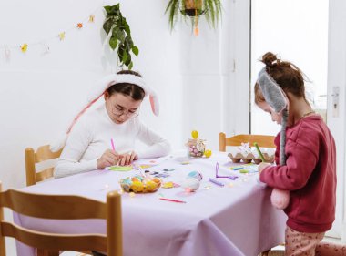Tavşan bantlı iki beyaz kız kardeşin portresi, bağımsız ve birlikte hazırlanıyorlar, keçeli kalemlerle Paskalya yumurtalarını boyuyorlar, bir bahar günü mutfakta açık eflatun kağıt masa örtüsüyle masada oturuyorlar, yandan bana bakıyorlar.